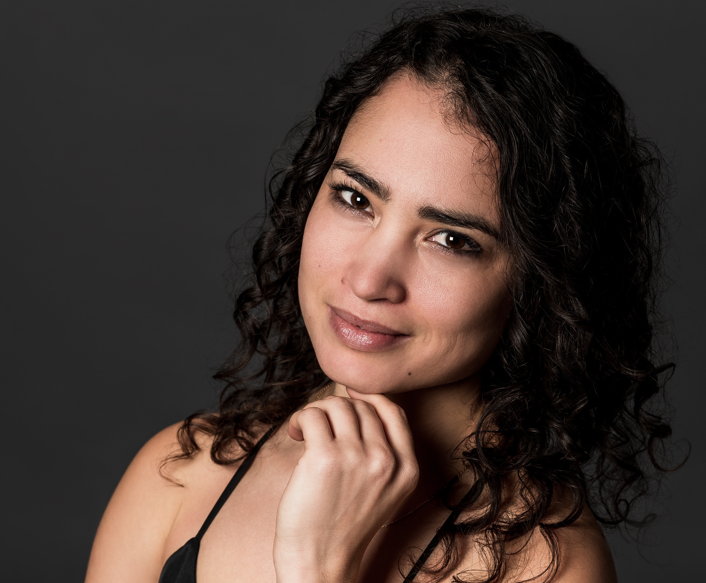 Close-up of a woman with dark curly hair, resting her chin on her hands against a dark gray background.