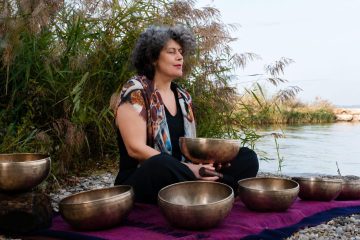 Woman meditating by a lakeshore with Tibetan singing bowls arranged on a purple mat