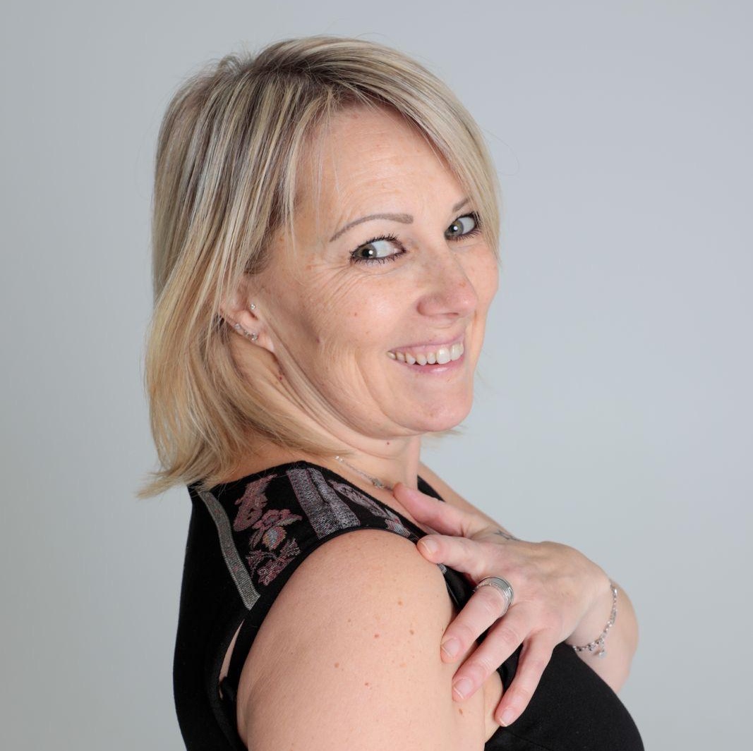 Smiling blonde woman in a black sleeveless top, looking over her shoulder at the camera against a light gray background.